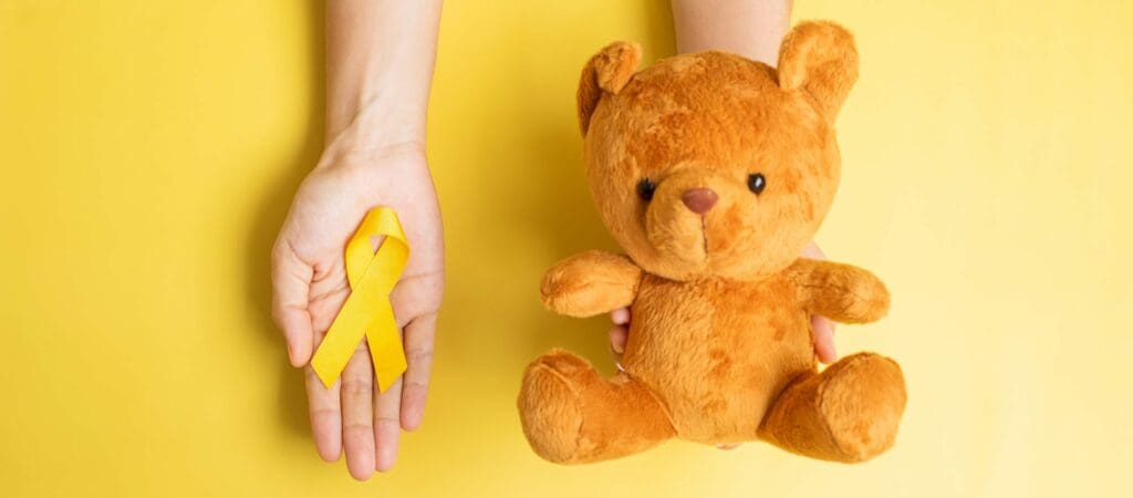 hand holding Yellow Ribbon and Bear doll on yellow background for supporting kid living and illness. September Childhood Cancer Awareness month and World cancer day concept