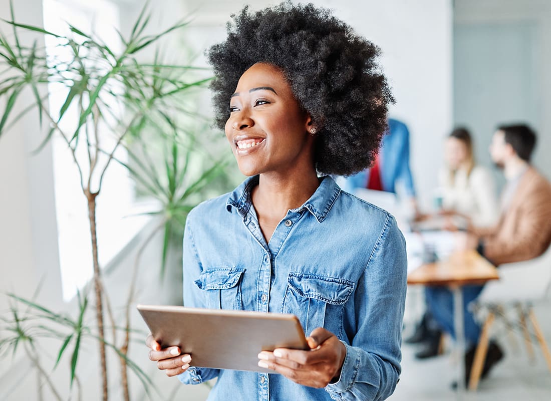 Employee Benefits - Portrait of a Cheerful Young Employee Standing in a Bright Conference Room Next to a Plant While Holding a Tablet in her Hands