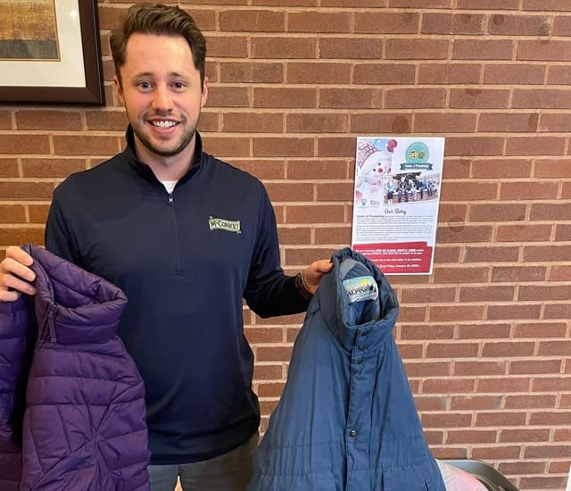Social Responsibility - Smiling Young McConkey Employee Holding Up Two Winter Jackets During a Coat Drive Event