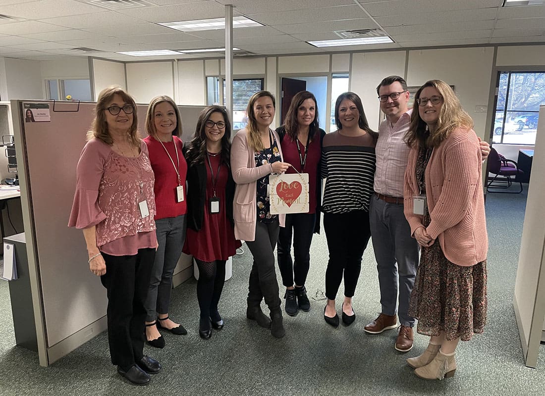 Contact - Portrait of a Small Group of Smiling McConkey Employees Standing in the Office While Wearing Pink and Red with One Employee Holding Up a Sign with a Heart