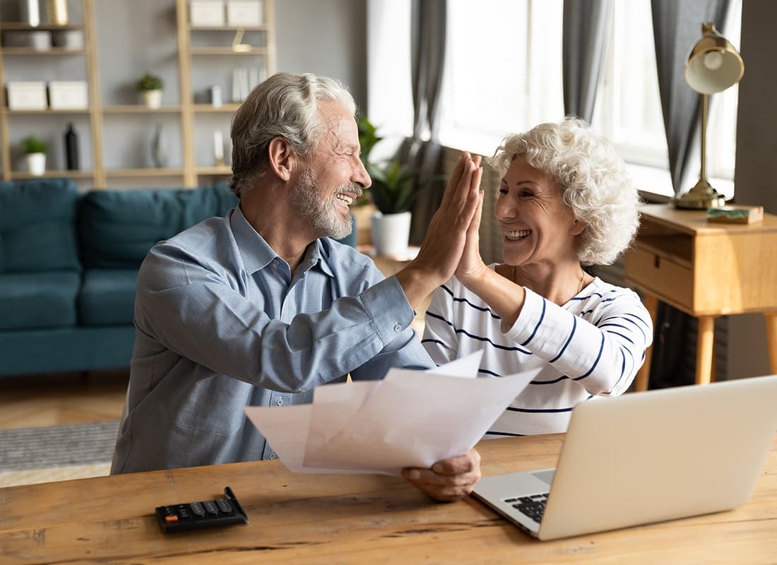 Personal Financial Protection - Overjoyed Elderly Couple Holding Paperwork While Sitting at a Desk with a Laptop in the Living Room Giving Each Other High Five