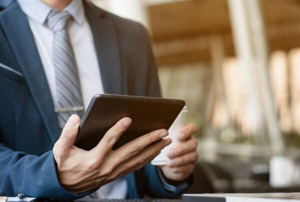 Blog - Business Man Looking at Tablet in Front of Open Laptop in a Cafe