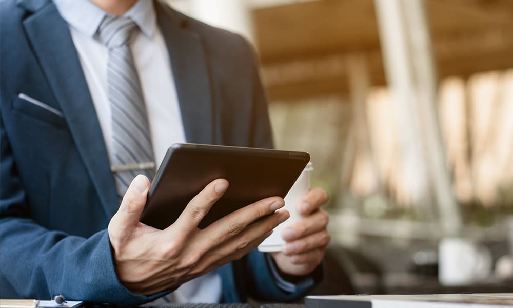 Blog - Business Man Looking at Tablet in Front of Open Laptop in a Cafe