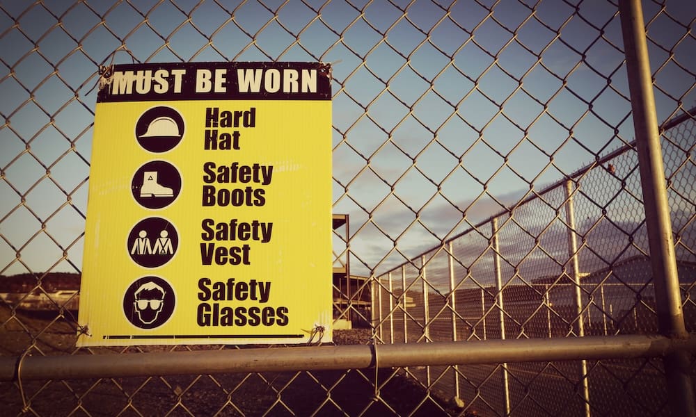 5 Essential Strategies for Contractors to Reduce Their Total Cost of Risk - Sign on a chainlink fence stating hard hat, safety boots, safety vest and safety glasses must be worn on the job site