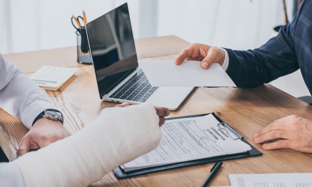 Understanding How Your Workers’ Compensation Rate is Calculated - Insurance agent at a desk with a workers comp claim on it handing a work in a cast a envelope